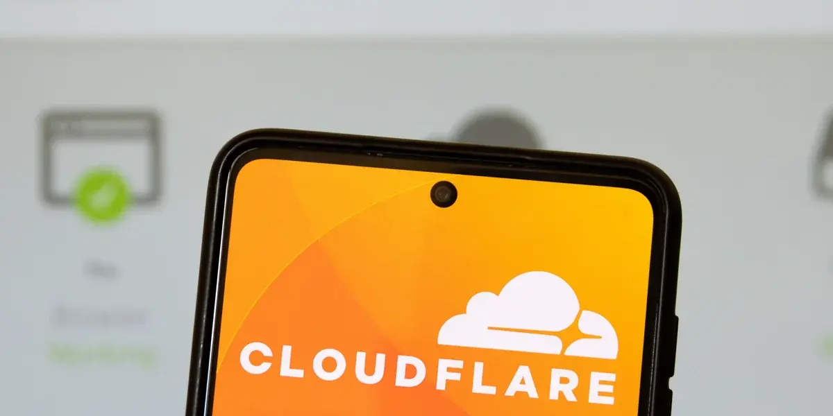 Telefon cu sigla Cloudflare pe ecran, în fața unei pagini de stare de server, neclară, cu o bifă verde