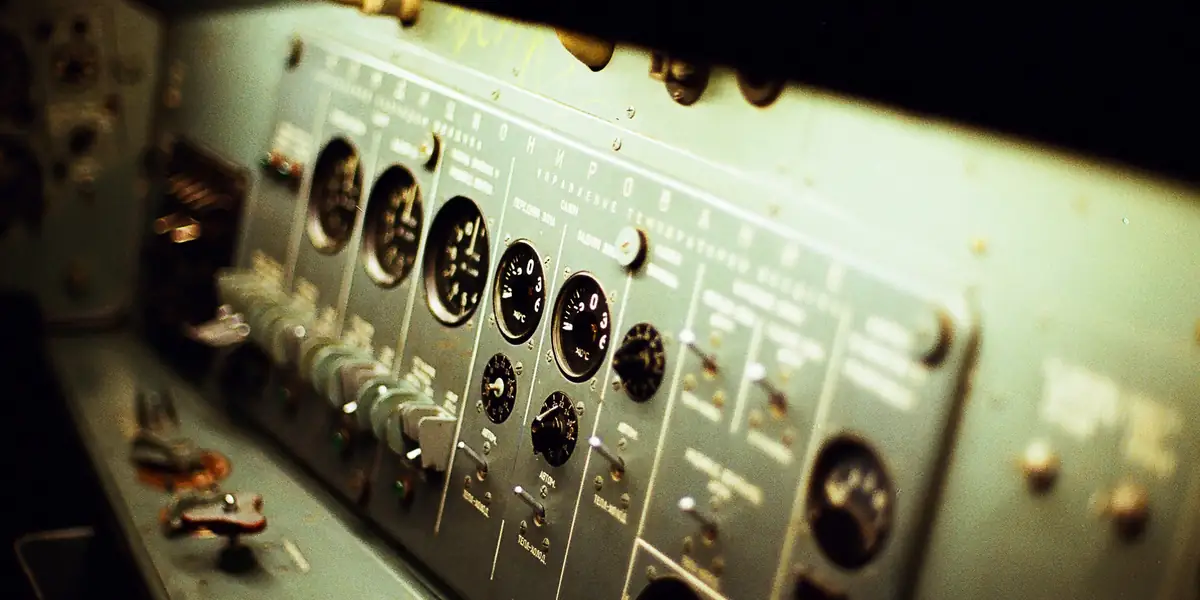 Metal control console shot at an angle, a row of identical toggle switches, circular dials, and a selector labelled AVTOM. under warm light