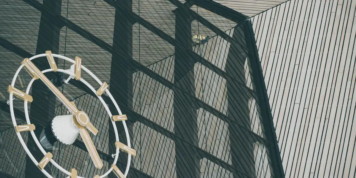Large white clock face with no numerals, hanging behind a dark steel-framed glass wall, photographed from below