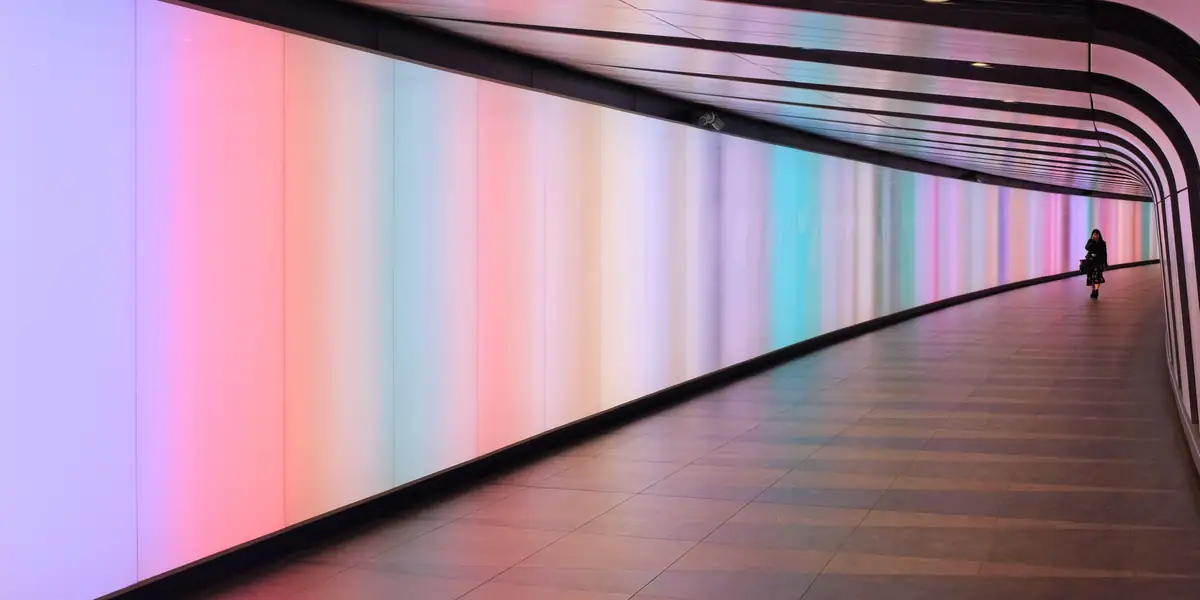 Curved walkway with an illuminated wall fading from pink through to pale blue, timber floor, one distant figure in silhouette