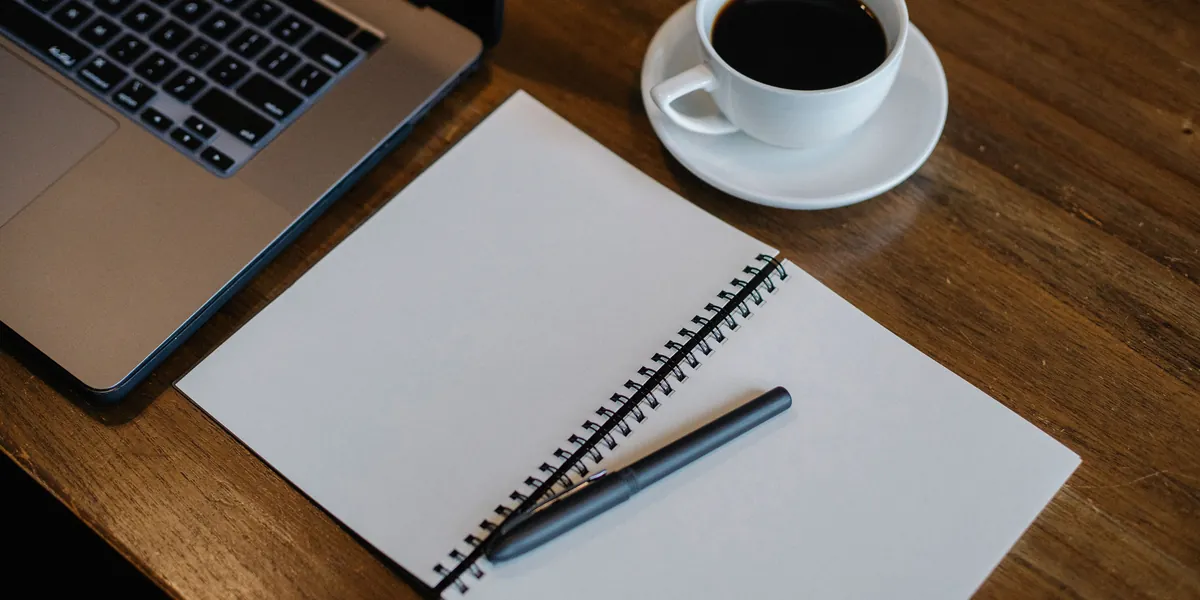 Quaderno aperto e vuoto con una penna sopra, una tazza di caffè e il bordo di un portatile su un tavolo di legno scuro, visti dall'alto