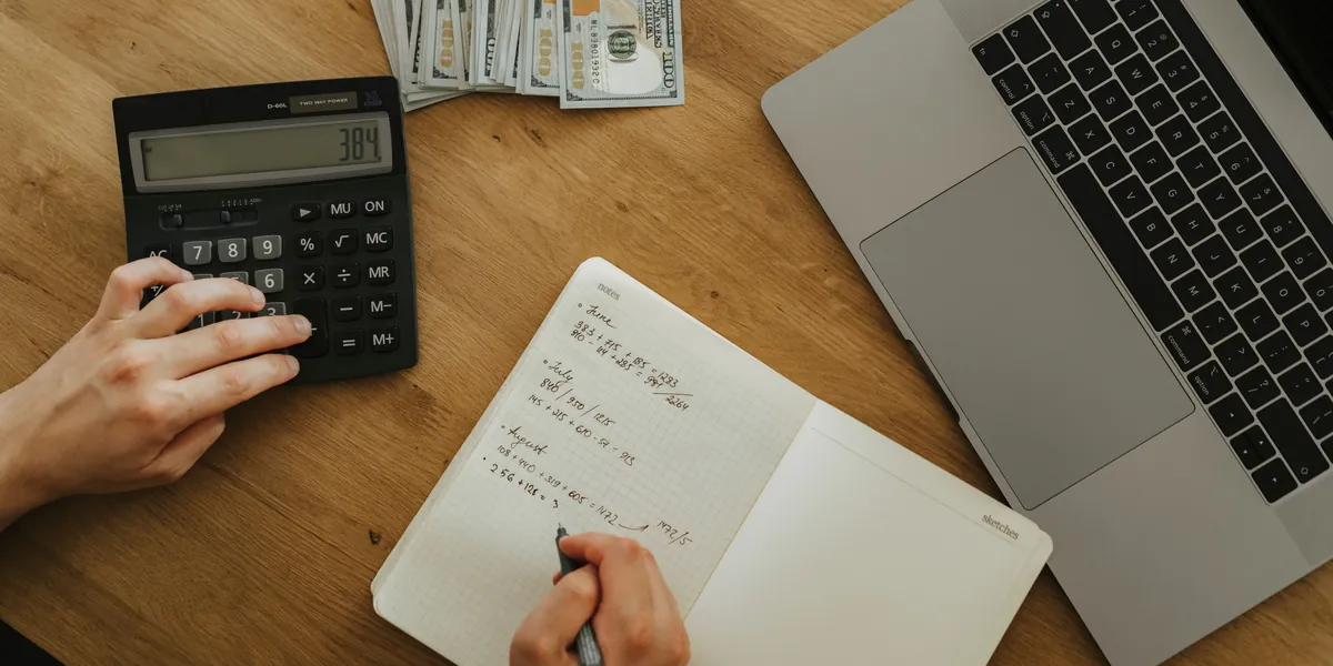 Mani che digitano su una calcolatrice nera accanto a un quaderno con i conti mensili scritti a mano, un portatile e alcune banconote, visti dall'alto