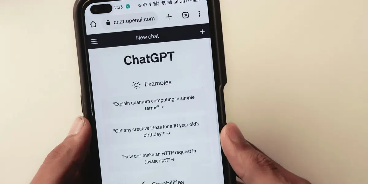 Două mâini țin un telefon în fața unui perete crem, browserul deschis pe chat.openai.com arată ecranul New chat cu titlul ChatGPT și exemple de întrebări, printre care Explain quantum computing in simple terms și How do I make an HTTP request in Javascript