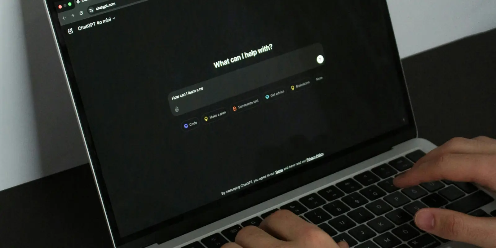 Două mâini care tastează pe tastatura unui laptop, ecranul arată chatgpt.com cu selectorul de model pe ChatGPT 4o mini și caseta de întrebare What can I help with, iar sub casetă rândul gri By messaging ChatGPT, you agree to our Terms and have read our Privacy Policy