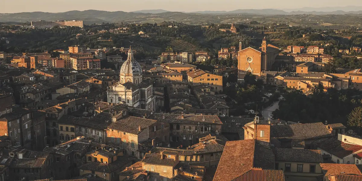Terracotta rooftops and church towers of a Tuscan hill town seen from above in warm late-afternoon light, hills receding into haze