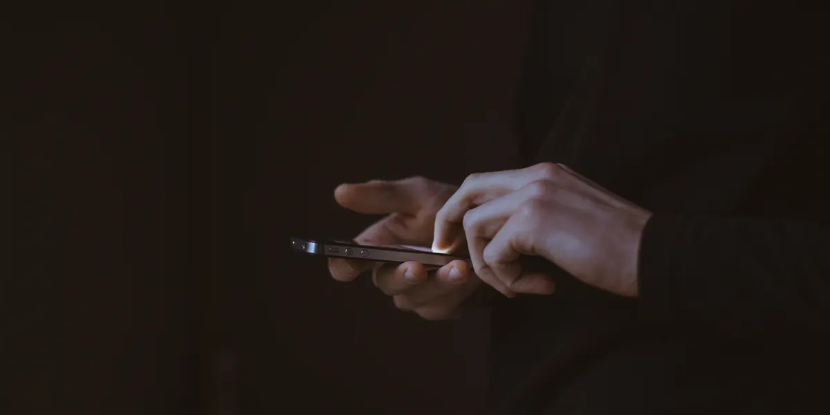 A hand holding a phone in near darkness, thumb resting on the screen, the rest of the frame almost entirely black