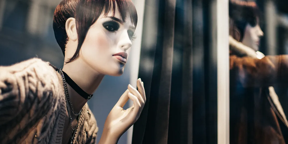 A shop-window mannequin with a brown bob and thin necklace seen through glass, street reflections across the pane and a second mannequin behind