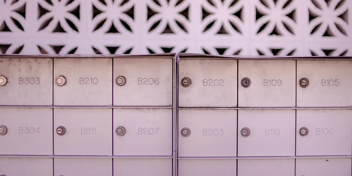 Two banks of pale metal post office boxes with engraved numbers B303, B210, B206, B202, B109, B105 on the upper row and B304, B211, B207, B203, B110, B106 below, under a decorative breeze-block wall
