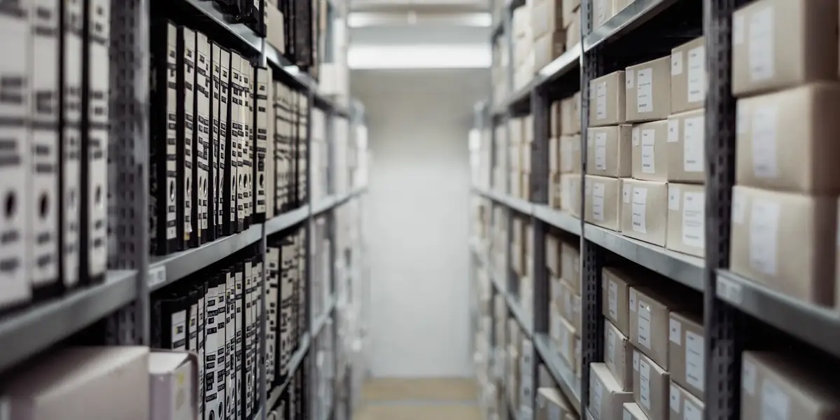 Long archive aisle with grey metal shelving holding hundreds of identical white and black box files stacked to the ceiling, labels blurred, corridor receding into pale light