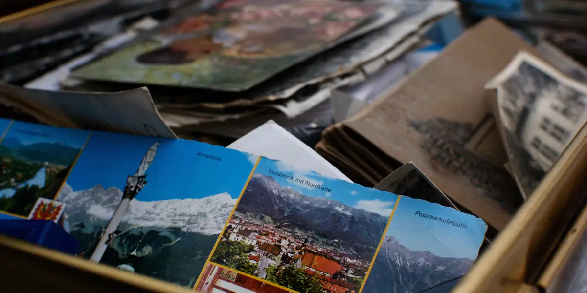 Open cardboard box of vintage Austrian tourist postcards, the top one showing Innsbruck mit Nordkette with insets labelled Annasaeule, Patscherkofelbahn and Hofkirche, older brown prints stacked behind