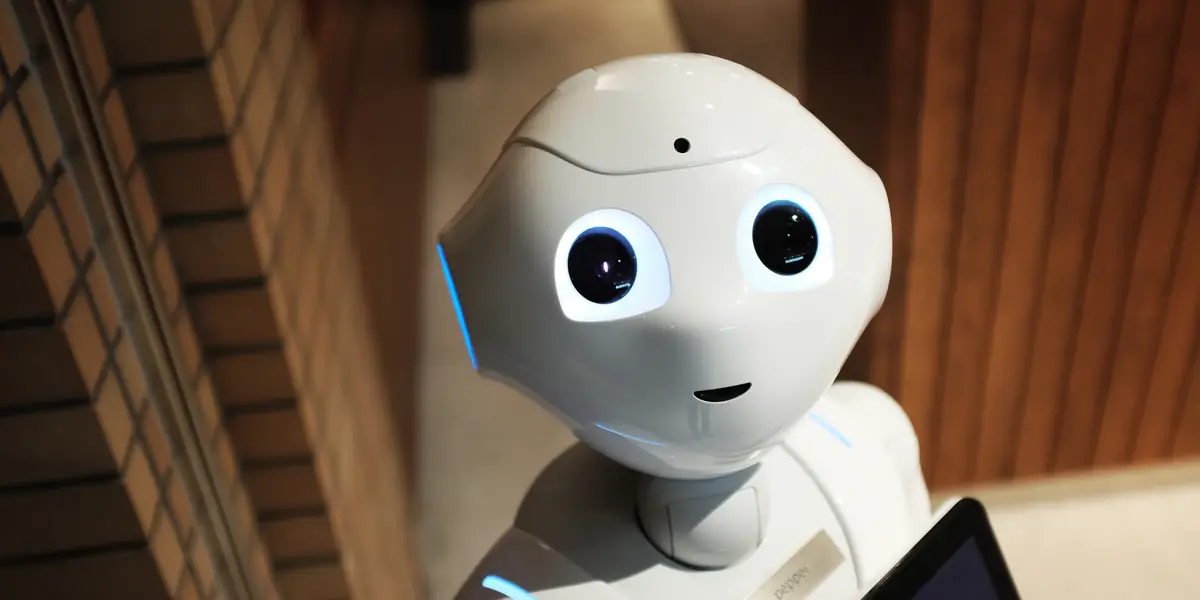 Robot umanoid alb cu ochi mari negri și o plăcuță pe piept pe care scrie pepper, fotografiat lângă un perete de lemn