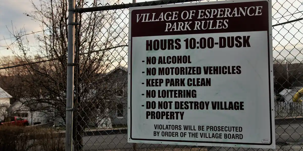 White sign bolted to a chain-link fence reading VILLAGE OF ESPERANCE PARK RULES, HOURS 10:00-DUSK, followed by five prohibitions and the line VIOLATORS WILL BE PROSECUTED BY ORDER OF THE VILLAGE BOARD