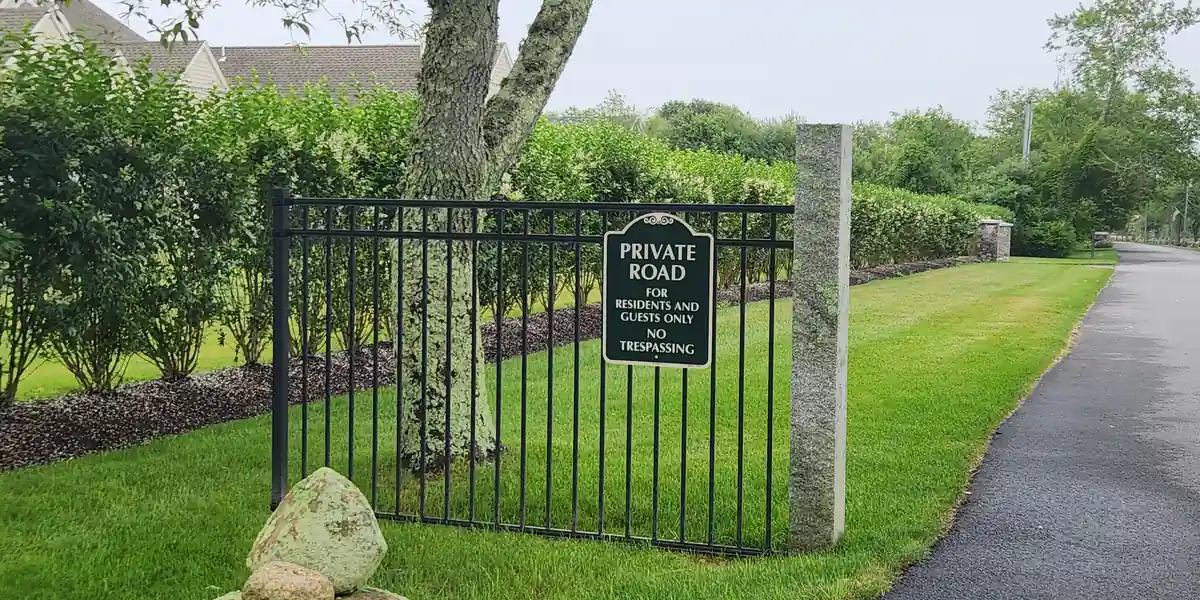 Short wrought-iron fence carrying an oval plaque that reads PRIVATE ROAD, FOR RESIDENTS AND GUESTS ONLY, NO TRESPASSING, with the tarmac path running past it unobstructed on the right