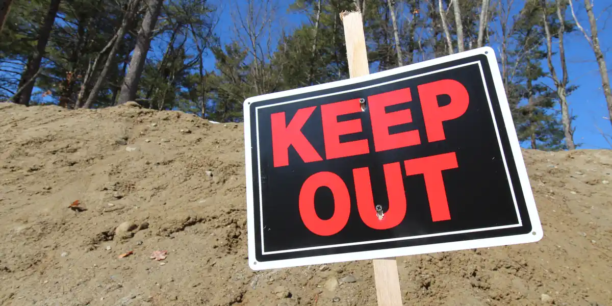 Black metal plate reading KEEP OUT in large red letters, fixed crookedly to a wooden stake driven into a mound of earth, trees and blue sky behind