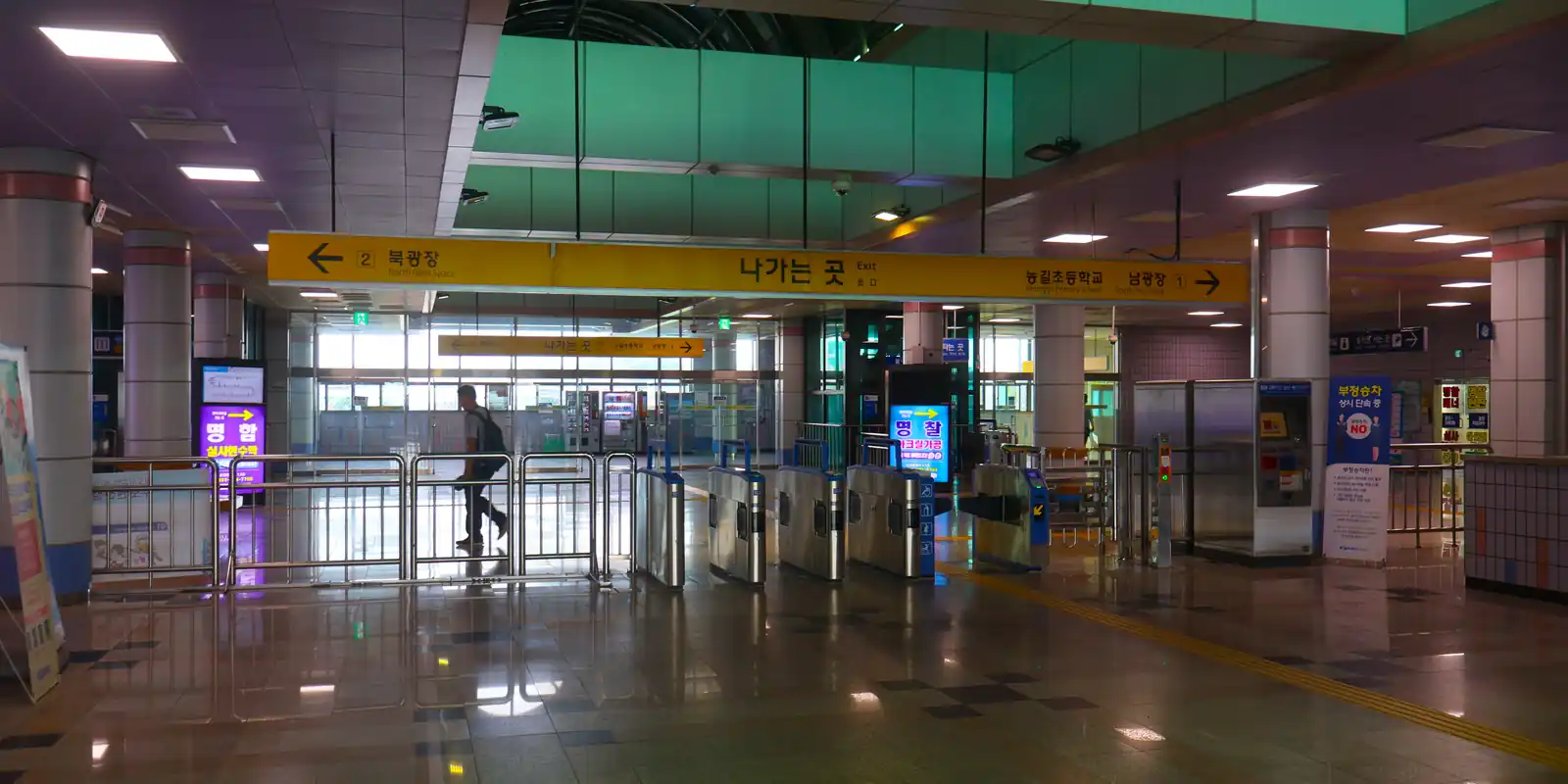 Empty underground station concourse with six ticket gates in a row beneath a yellow wayfinding sign, one passenger walking through them