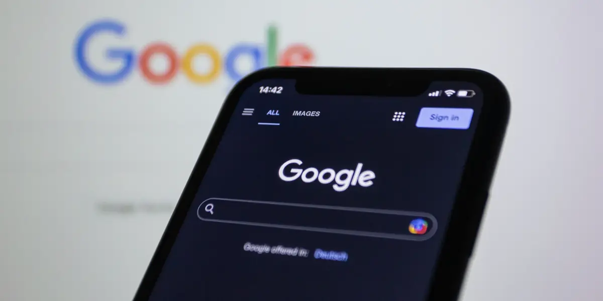Telefon cu aplicația Google deschisă, ținut în fața unui monitor pe care se vede aceeași pagină de căutare