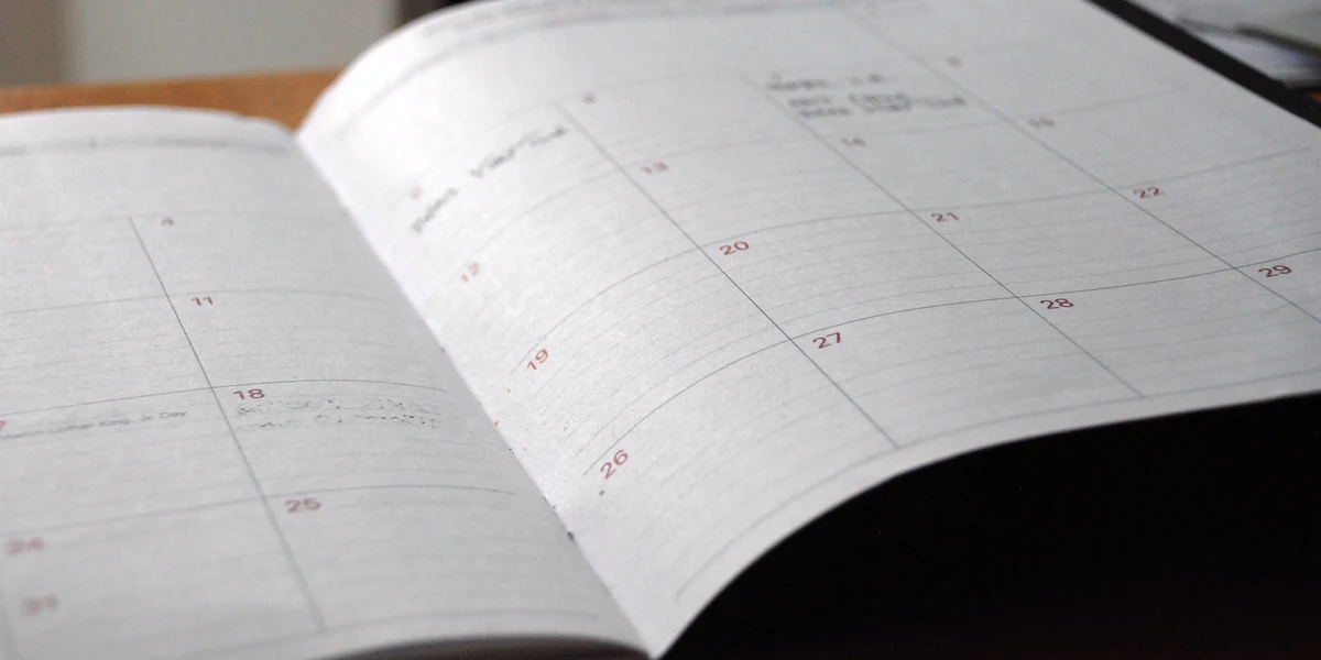 Open monthly planner on a wooden table showing the days 11, 18 and 25 on the left page and 13, 19, 20, 21, 22, 26, 27, 28, 29 on the right, handwritten notes illegible