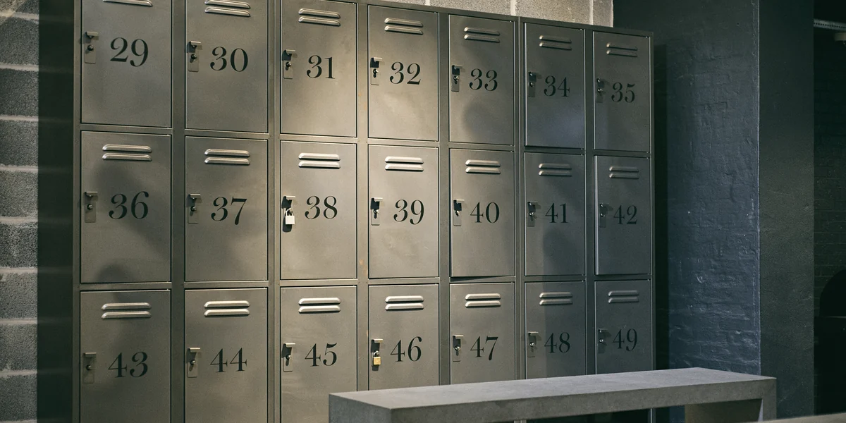 Three rows of grey metal lockers numbered 29 to 49, with latches and two padlocks fitted, against a brick wall with a concrete bench in front
