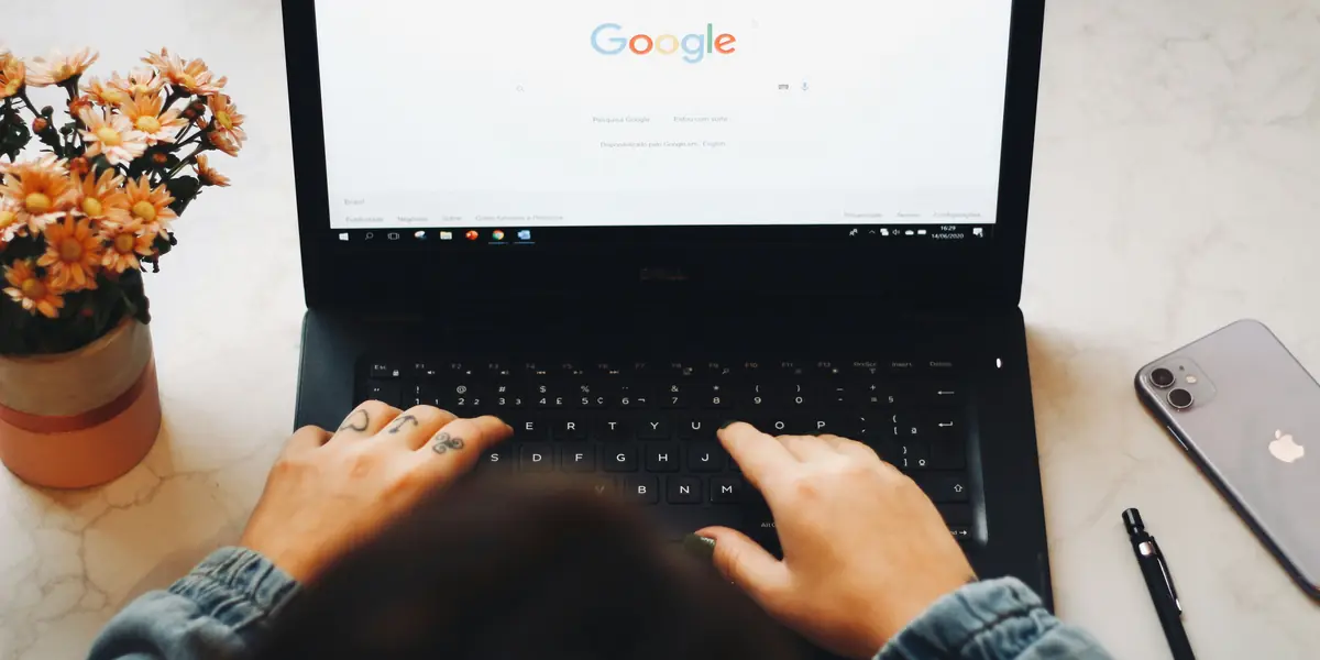 Vedere de sus peste un birou de marmură: două mâini tastează pe un laptop deschis la pagina de pornire Google, cu un telefon argintiu, un stilou negru și un ghiveci cu flori portocalii în jur