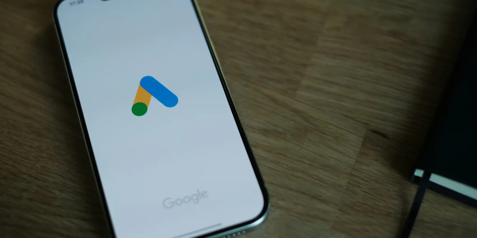 Un telefon așezat cu ecranul în sus pe un birou de lemn, arătând sigla Google Ads — triunghiul albastru cu punct verde și bară galbenă — pe fundal alb, cu cuvântul Google dedesubt, lângă coperta unui carnet negru
