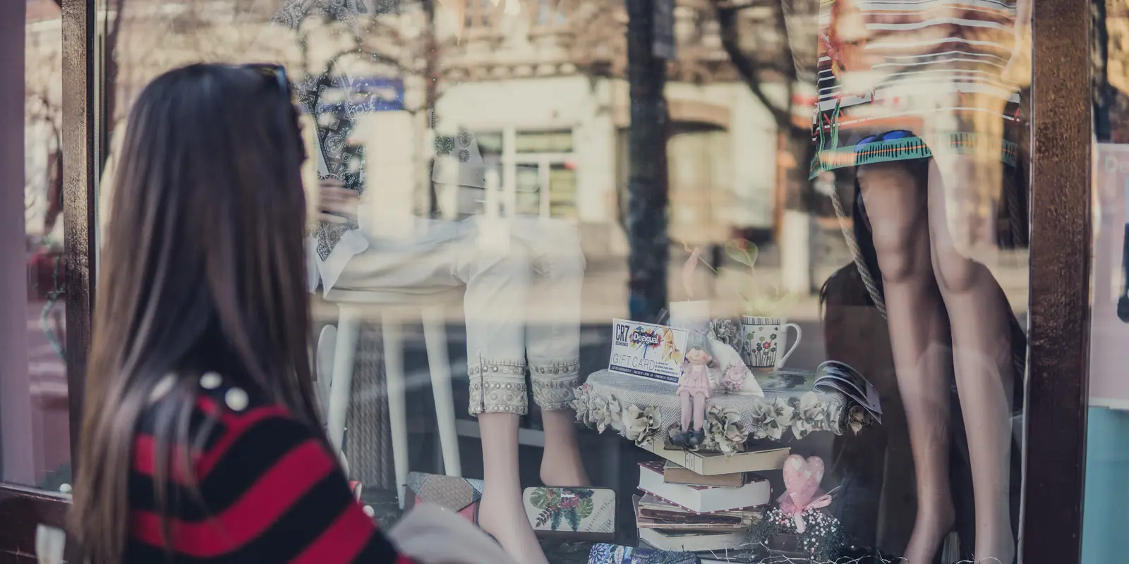 Una passante con maglione a righe rosse e nere si ferma sul marciapiede davanti alla vetrina di una boutique, tra manichini, tazze e libri impilati