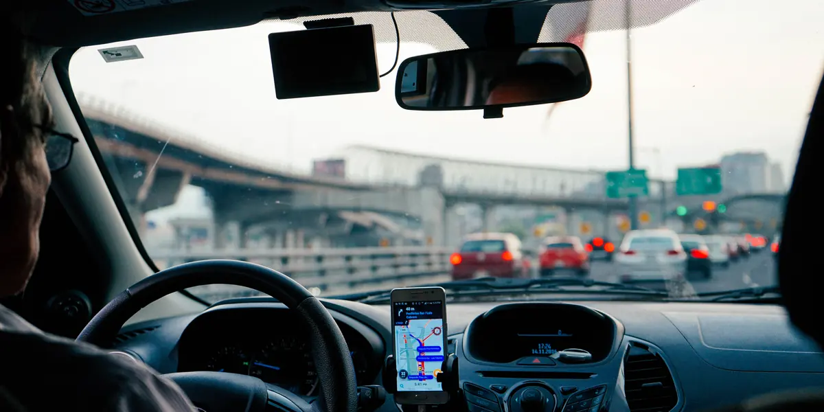 Vista dal posto di guida: smartphone fissato al cruscotto con una mappa di navigazione attiva, traffico intenso davanti e un cavalcavia che attraversa la carreggiata