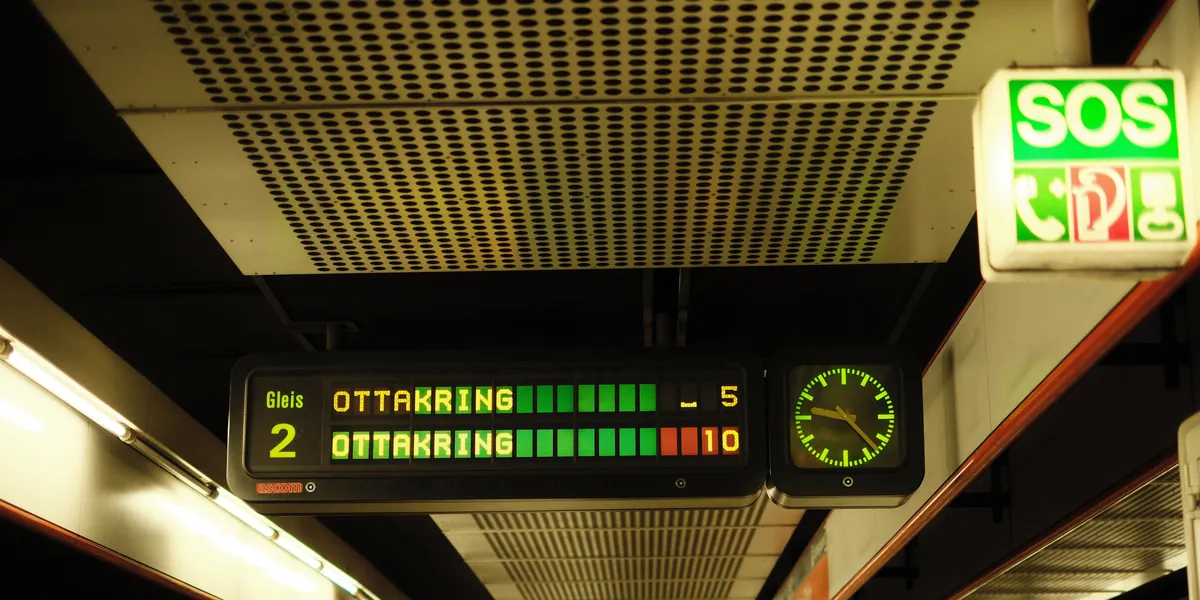 A Vienna underground platform display board in yellow and green characters showing the destination Ottakring and the waiting times 5 and 10 minutes, with an illuminated SOS sign beside it