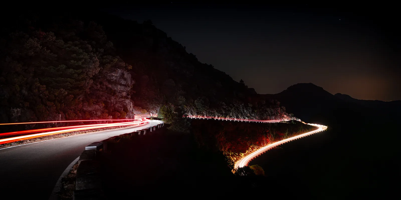 Long-exposure night photograph of a mountain road, with car headlights and tail lights drawn as continuous red and white ribbons along the curve