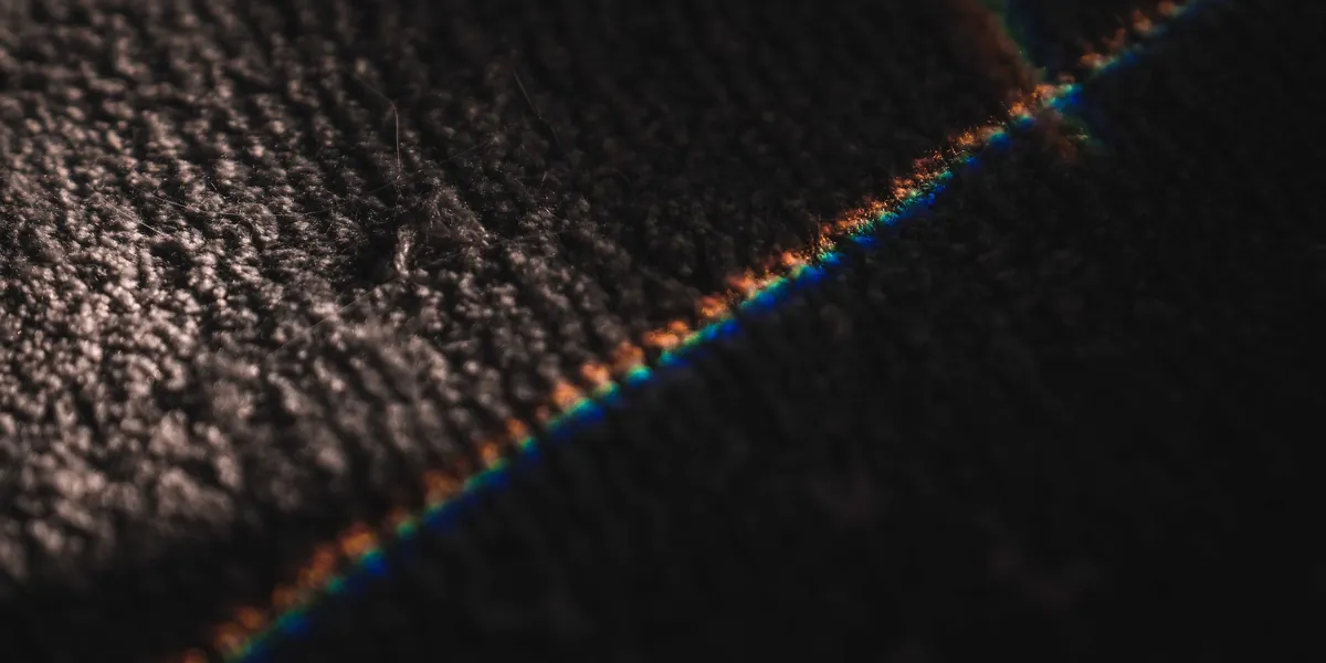 A dark, coarse surface filling the frame, crossed diagonally by a thin line of light split into rainbow colours, a few millimetres wide