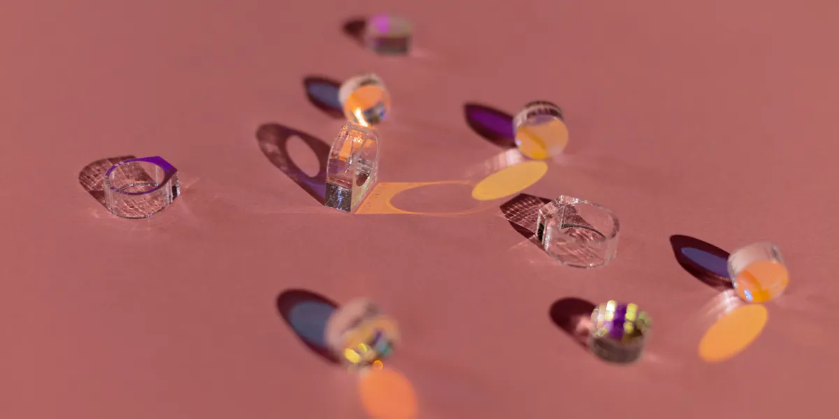 Small clear glass rings scattered across a dusty pink surface, each one throwing its own orange and violet coloured shadow beside it