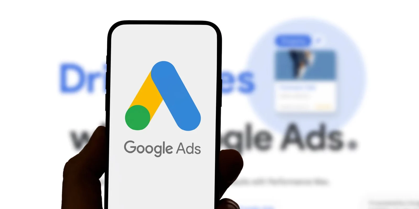 A hand holds a smartphone showing the Google Ads logo, with the “Drive sales with Google Ads” page behind it