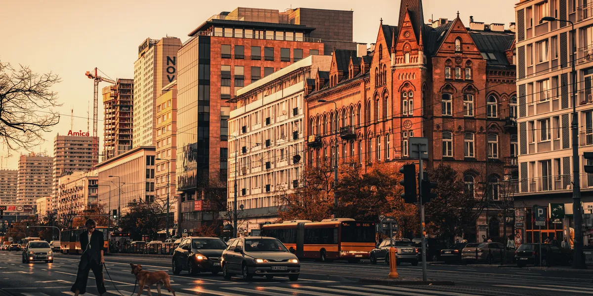 Una strada larga di Varsavia al tramonto, edifici storici e moderni affiancati, traffico e passanti nella luce bassa e dorata