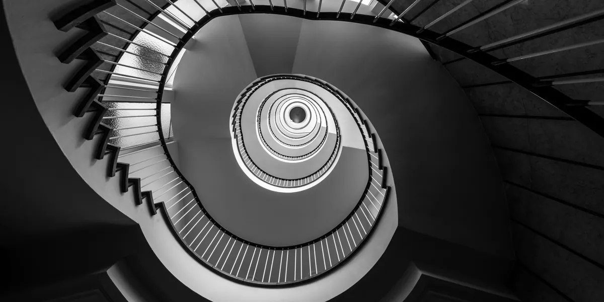Una scala a chiocciola fotografata dal basso, in bianco e nero, con i gradini che si rimpiccioliscono verso un punto luminoso al centro