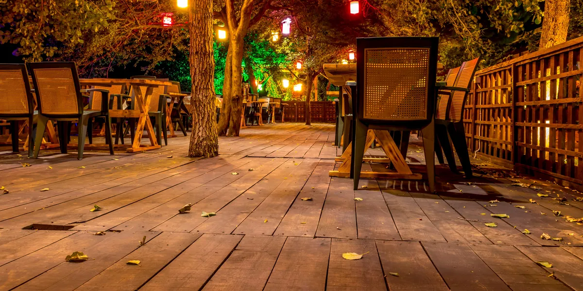 Un dehors di ristorante vuoto di sera, con tavoli e sedie di legno su un pavimento di assi, lampadine calde fra gli alberi e foglie cadute a terra