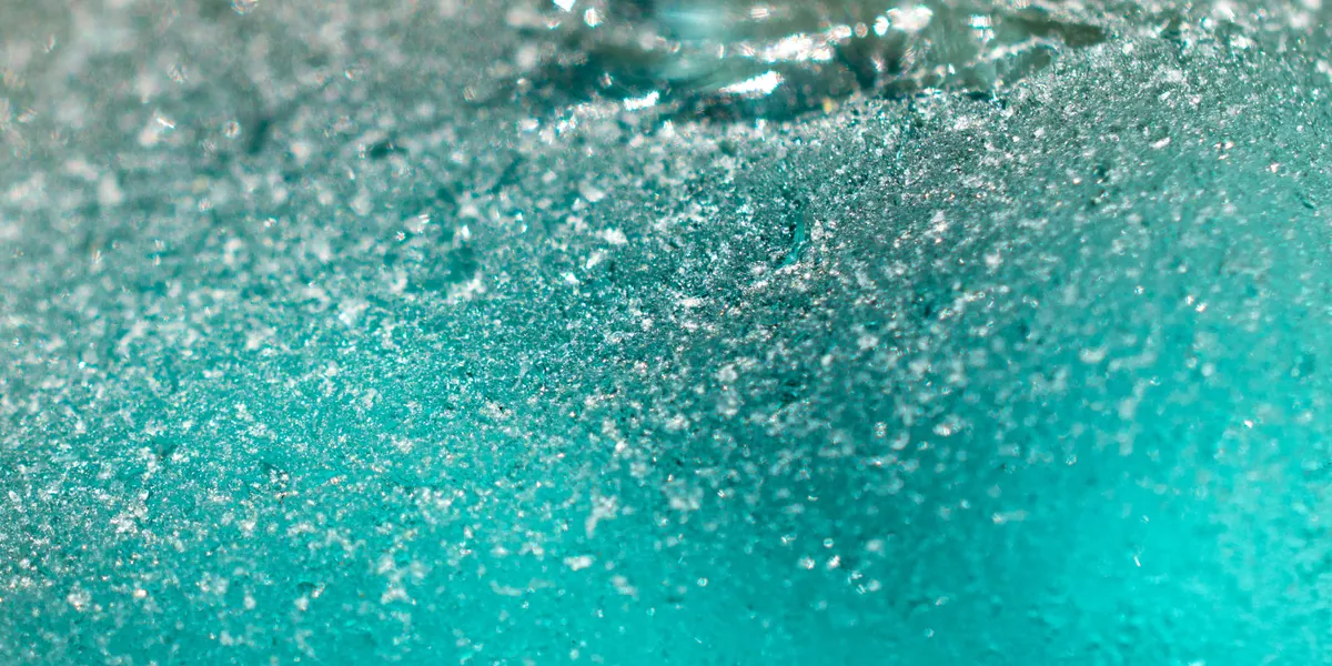 Primo piano macro su una superficie di vetro smerigliato color turchese, con granuli finissimi di brina che catturano la luce