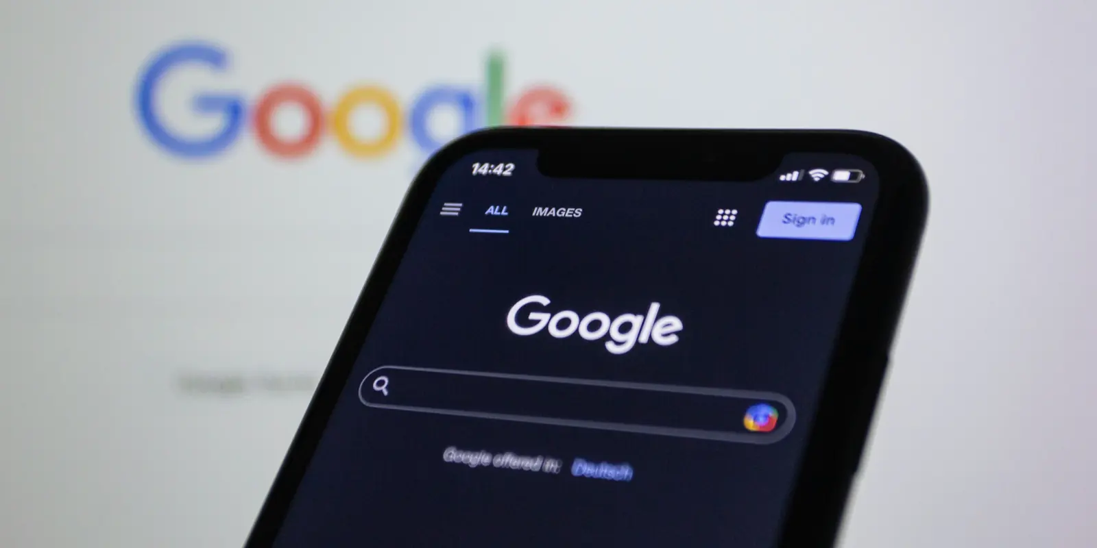 A phone showing Google search in dark mode, with the Google homepage on a screen behind it