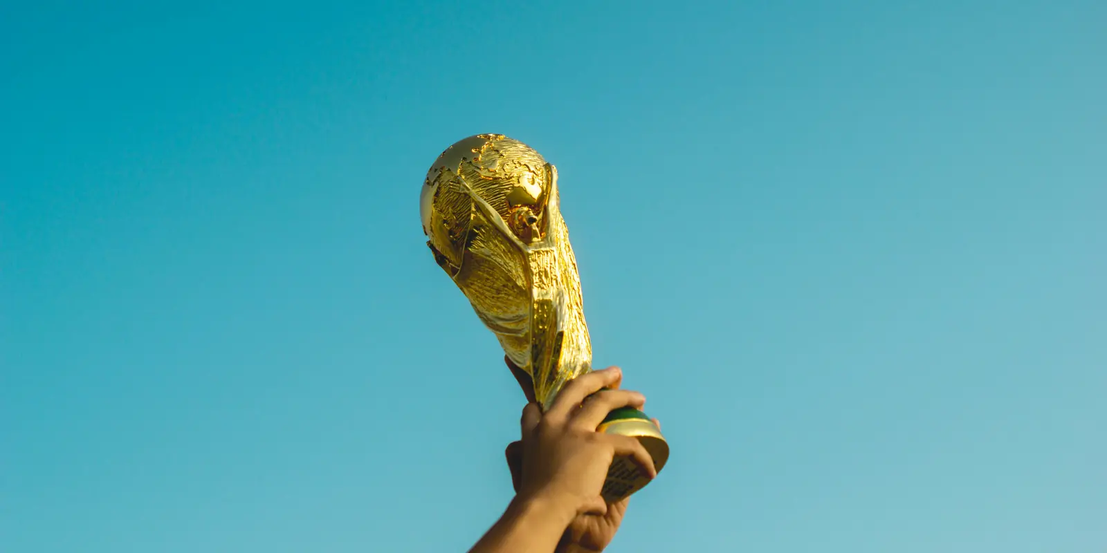 A hand raising a gold trophy against a clear blue sky