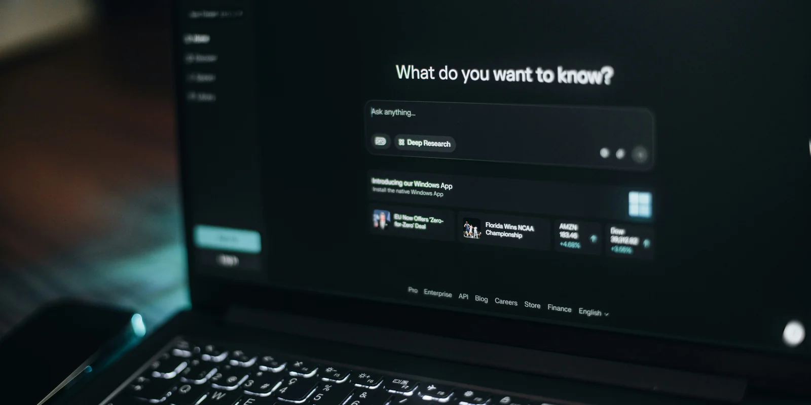 Un laptop într-o cameră întunecată arată un asistent de căutare AI cu întrebarea „What do you want to know?”