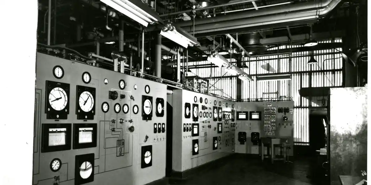 Fotografie alb-negru de arhivă dintr-o cameră de comandă industrială, cu zeci de cadrane rotunde și instrumente pătrate montate pe panouri de-a lungul întregului perete