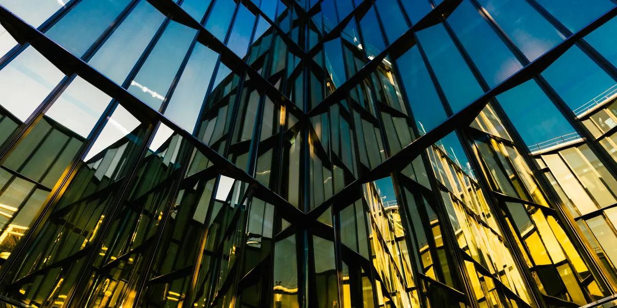 The glass facade of a modern building seen from below, its panels reflecting blue sky on one side and a golden light on the other