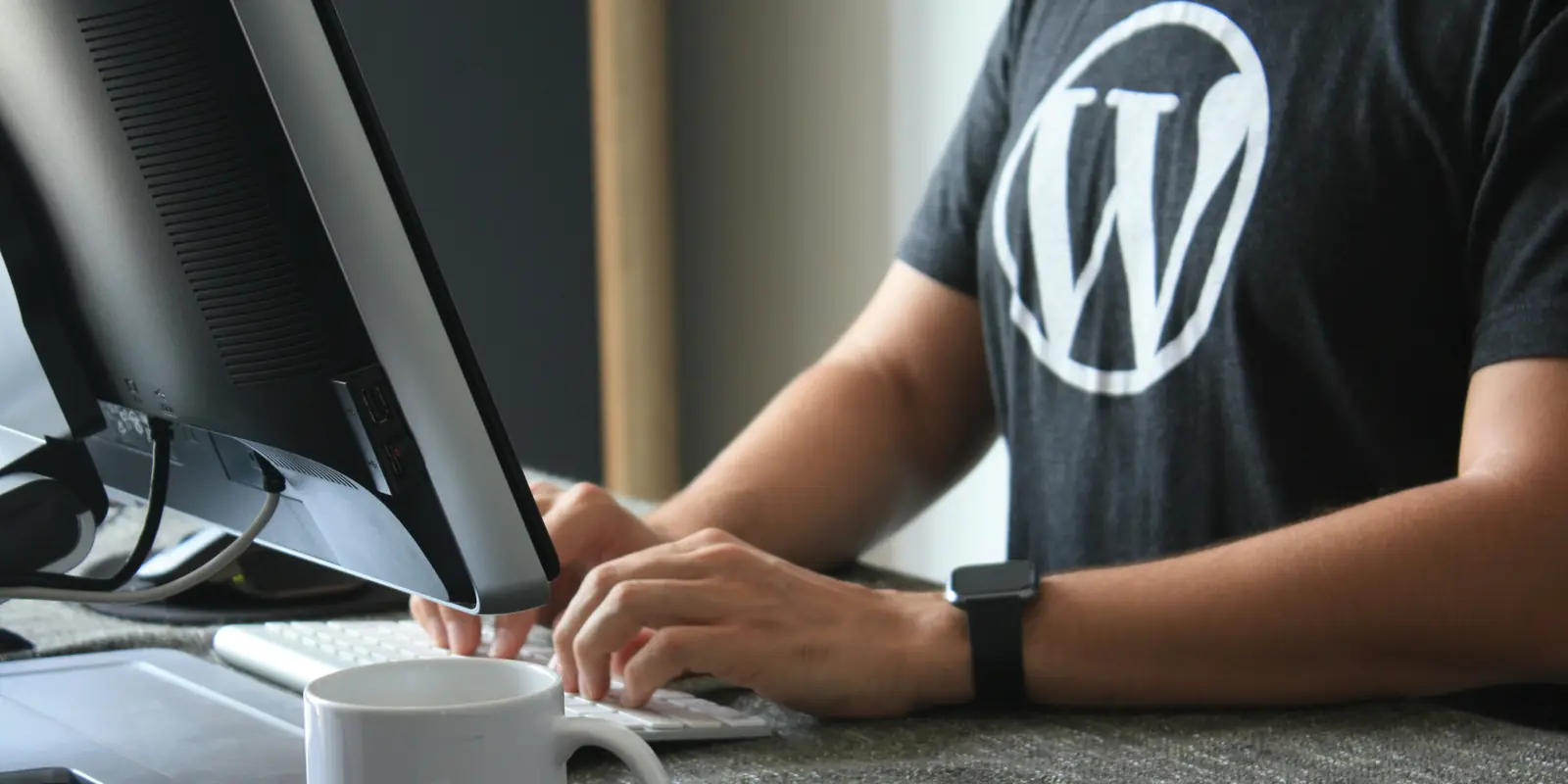 Person sitting at a desk typing on a keyboard in front of a monitor, wearing a dark T-shirt with the WordPress W logo, a white mug next to the keyboard