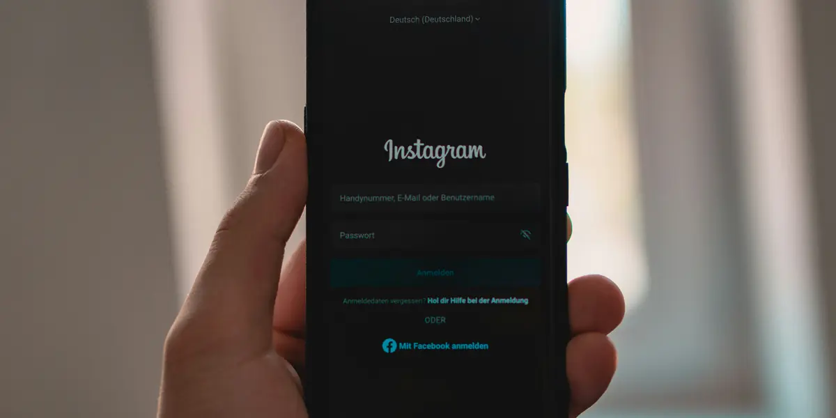 A hand holding a smartphone showing the Instagram login screen in German: fields for phone number or username and for the password, and below them a blue Mit Facebook anmelden button to sign in with a Facebook account
