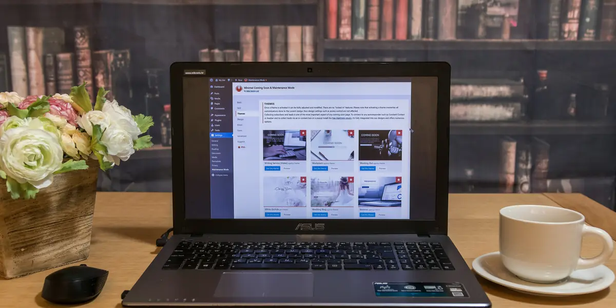 Un laptop deschis pe un birou de lemn deschis la culoare, lângă el o ceașcă albă de cafea și un ghiveci de lemn cu flori roz și albe, iar pe ecran panoul de administrare WordPress cu bara laterală întunecată, meniul Aspect deschis și ecranul temelor