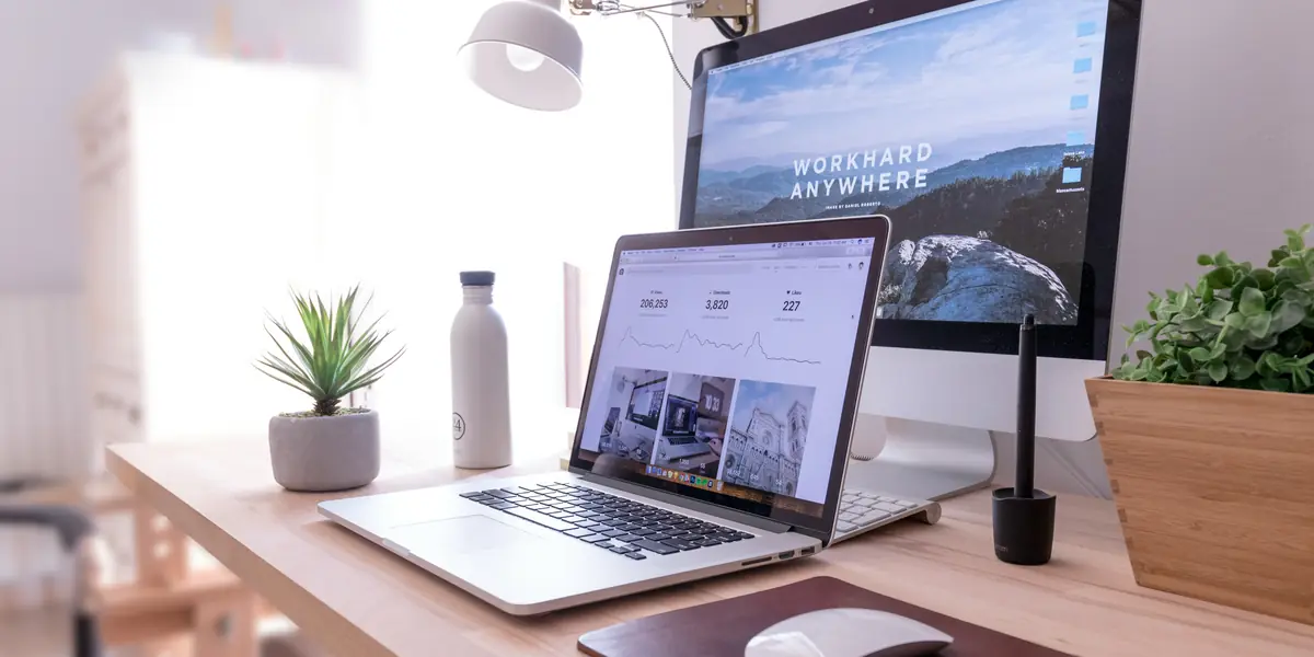 Birou luminos cu un iMac care afișează textul Work hard anywhere peste o fotografie de munte, în față un laptop deschis pe un panou de statistici cu cifrele 206.193, 3.820 și 227 deasupra a trei grafice, lângă o sticlă albă de metal și o plantă suculentă