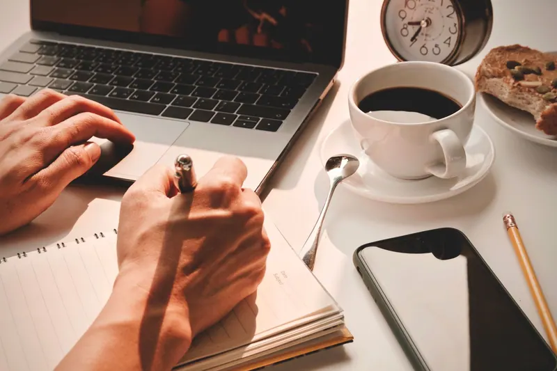 Hands writing a plan in a notebook, next to a laptop and a coffee